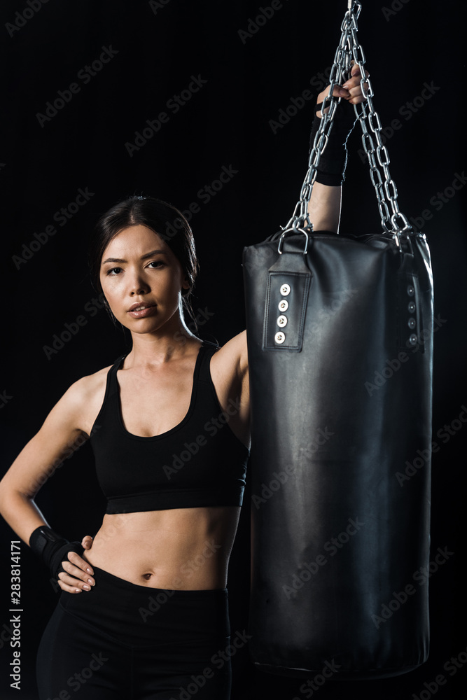 attractive girl standing with hand in hip near punching bag isolated on black