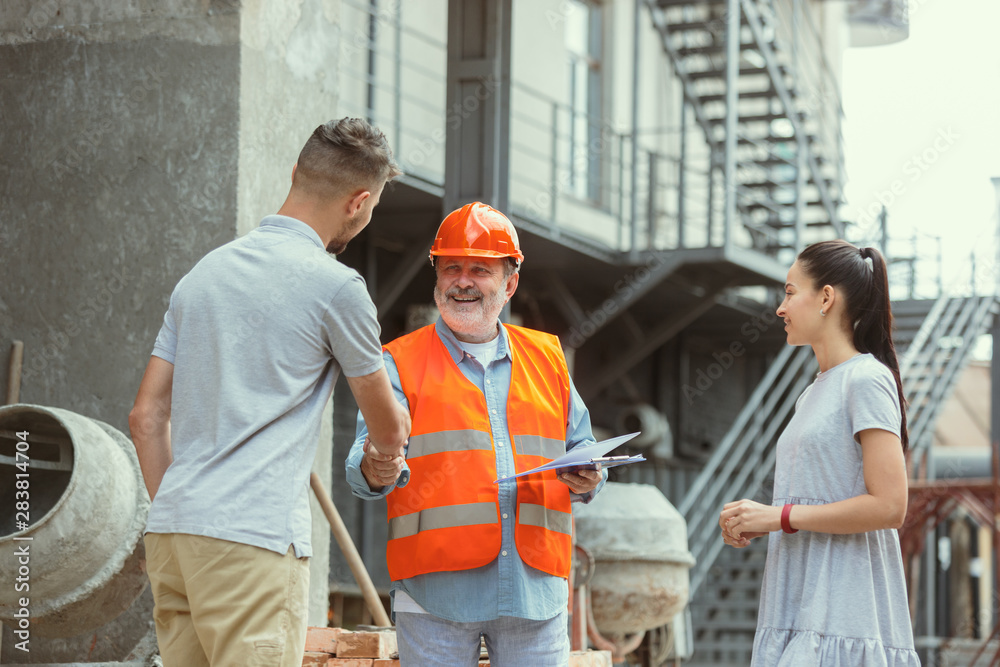 Foreman or achitect engineer shows future house, office or store design plans to a young couple. Meeting at the construction site to talk about facade appearance, interior decoration, home layout.