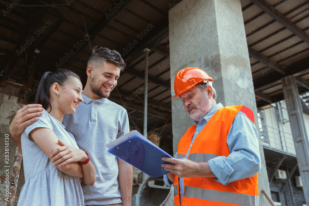 Foreman or achitect engineer shows future house, office or store design plans to a young couple. Meeting at the construction site to talk about facade appearance, interior decoration, home layout.