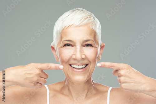 Beautiful Caucasian  smiling senior woman with short grey hair pointing at her teeth and looking at camera. Beauty photography. photo