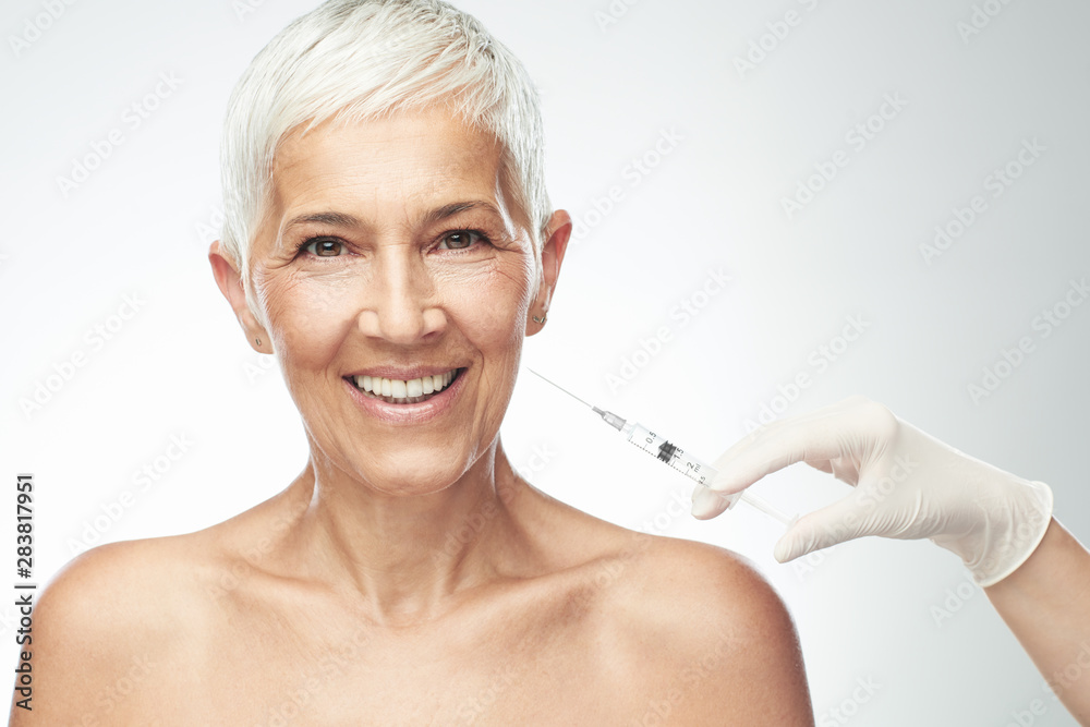 Beautiful smiling Caucasian senior woman standing in front of gray background and taking a shot of anti age product. Beauty photography.