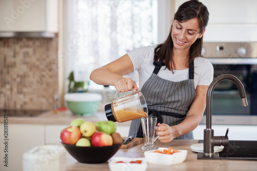 Beautiful smiling Caucasian woman in apron standing in kitchen and pouring fresh smoothie into glass.