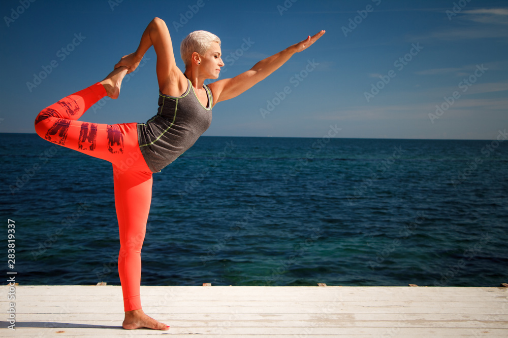 Obraz premium Adult blond woman with short haircut practices yoga on the pier against the background of the sea and blue sky