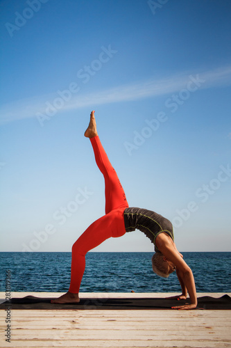 Adult blond woman with short haircut practices yoga on the pier against the background of the sea and blue sky © Sahaidachnyi Roman