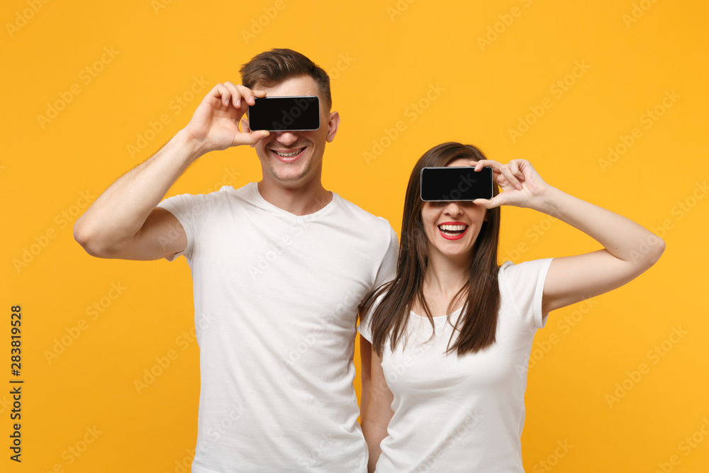 Young couple two friends guy girl in white t-shirts posing isolated on yellow orange background. People lifestyle concept. Mock up copy space. Covering eyes with mobile phone with blank empty screen. Young couple two friends guy girl in white t-shirts posing isolated on yellow orange background. People lifestyle concept. Mock up copy space. Covering eyes with mobile phone with blank empty screen.