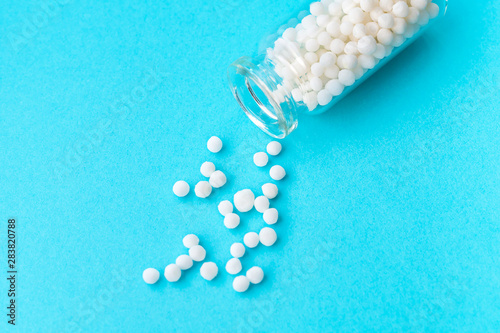 Homeopathic globules in glass bottle on pastel blue background. The globules are scattered from bottle. Alternative homeopathy medicine herbs, healthcare and pills concept. Selective focus photo