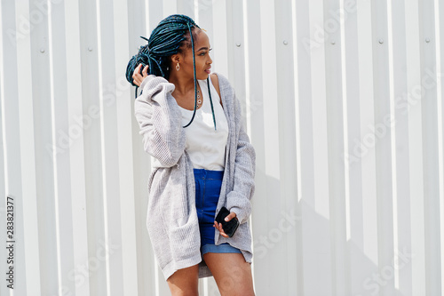 African woman with dreads hair, in jeans shorts  posed against white steel wall with mobile phone in hand. © AS Photo Family