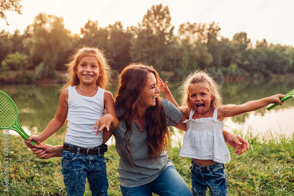 Family having fun after playing badminton by summer river at sunset. Mother laughing and grimacing with daughters Family having fun after playing badminton by summer river at sunset. Mother laughing and grimacing with daughters