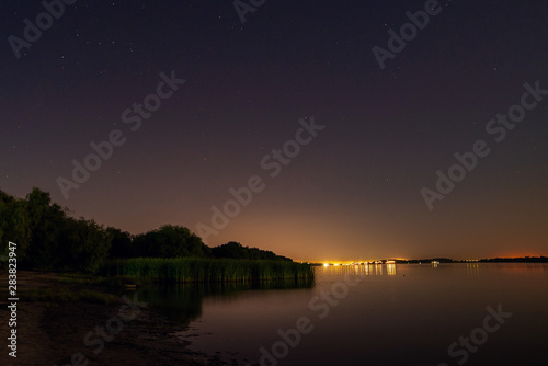 Lake or river sand shore with trees and dark blue starry sky and city light on background. Tranquil nature night landscape
