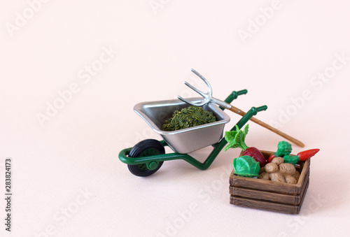 Realistic figures of farm or garden tools: iron wheelbarrow with green knob, pitchfork, hay, near wooden box of vegetables: potatoes, carrot, cabbage, beet on the light background