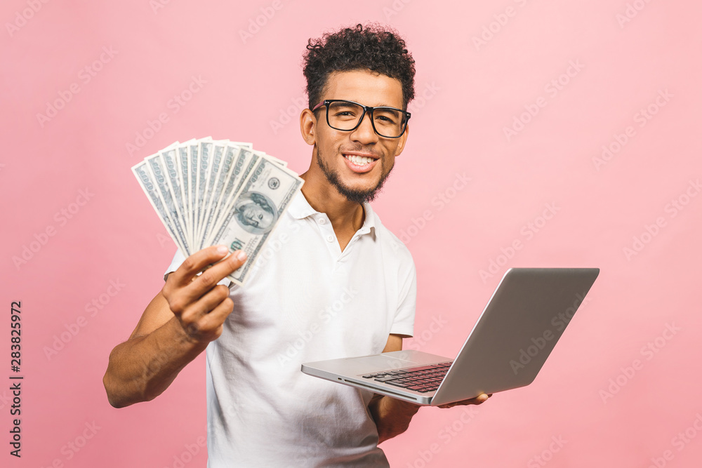 Closeup portrait, young successful african american business man making money from internet, holding cash in hand, laptop in another, isolated over pink background. Closeup portrait, young successful african american business man making money from internet, holding cash in hand, laptop in another, isolated over pink background.