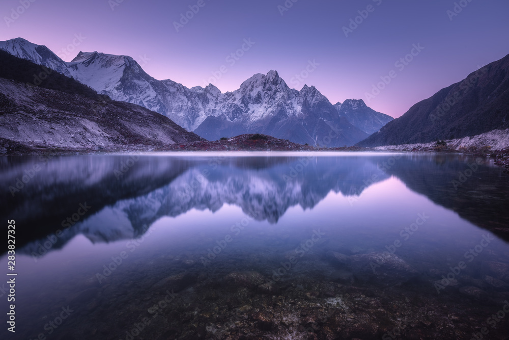 Mountain lake with perfect reflection at sunrise. Beautiful landscape with purple sky, snowy mountains, hills, fog over the lake at twilight in Nepal. Snow covered rocks is reflected in water. Nature