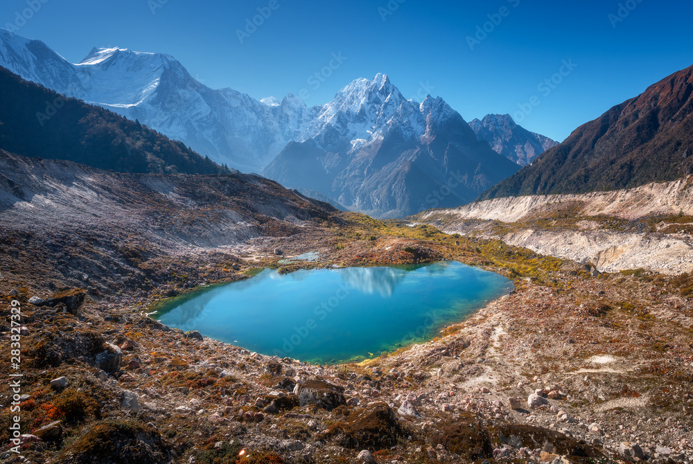 Small lake with blue water and sky reflection against beautiful snow covered mountains at sunny day in autumn. Landscape with lake, snowy rocks, hills, stones at sunrise. Himalayan mountains in Nepal Small lake with blue water and sky reflection against beautiful snow covered mountains at sunny day in autumn. Landscape with lake, snowy rocks, hills, stones at sunrise. Himalayan mountains in Nepal