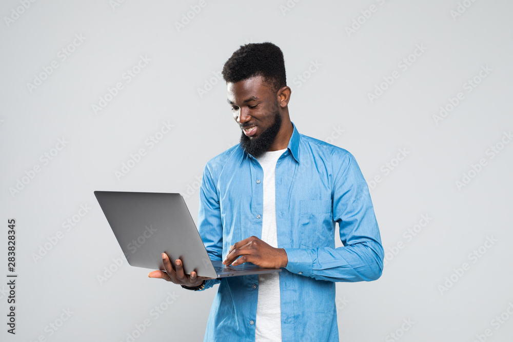 Young smiling african man standing and using laptop isolated over gray background