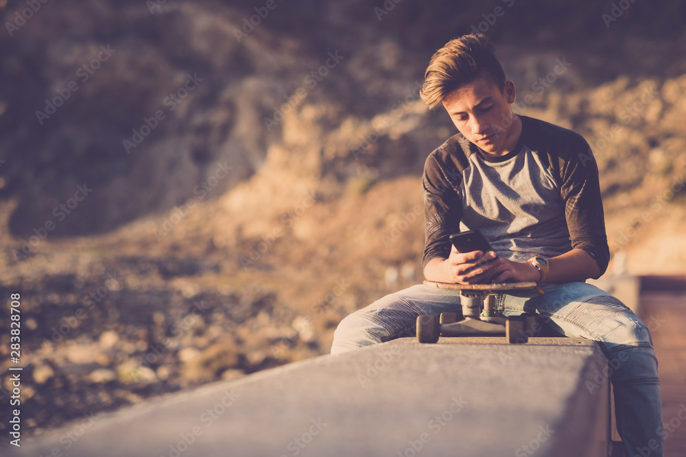 teenager at the beach sitting on a wall using his phones with his skateboard - boy wearing jeans focused on the social or playing video games