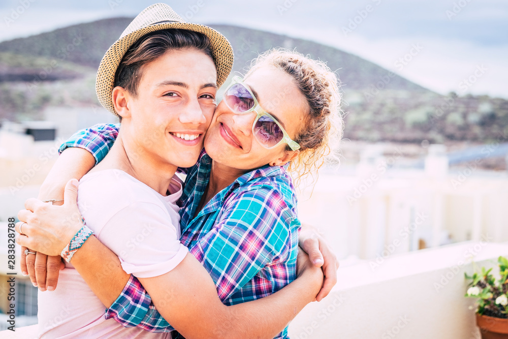beautiful couple of mum and son together hugged in the terrace of the home having fun and laughing - woman with sunglasses and teenager with a relationship - guy with a hat hapy and funny