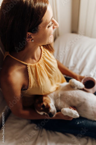 Top view of beautiful woman sitting on the bed looking out the window with her dog Jack Russell terrier lying in her legs and a cup of coffee at sunset