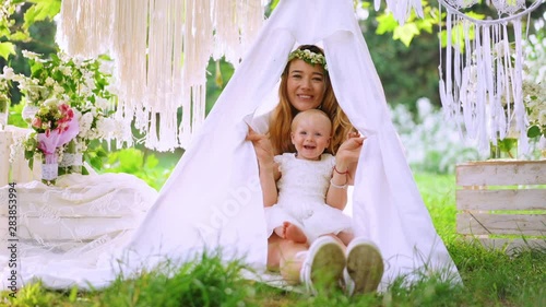 A baby with her mother hides in the wigvam on the green grass. Outside shooting, the garden on the background. Portrait. Party atmosphere. photo