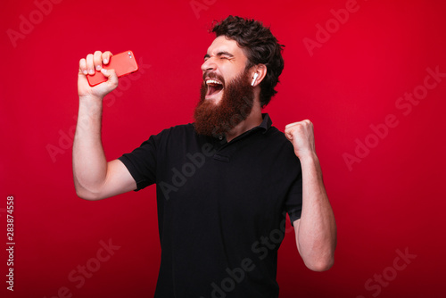 Photo of bearded guy, hoding mobile phone, using wireless earphones and enjoying music, over red bcakground photo