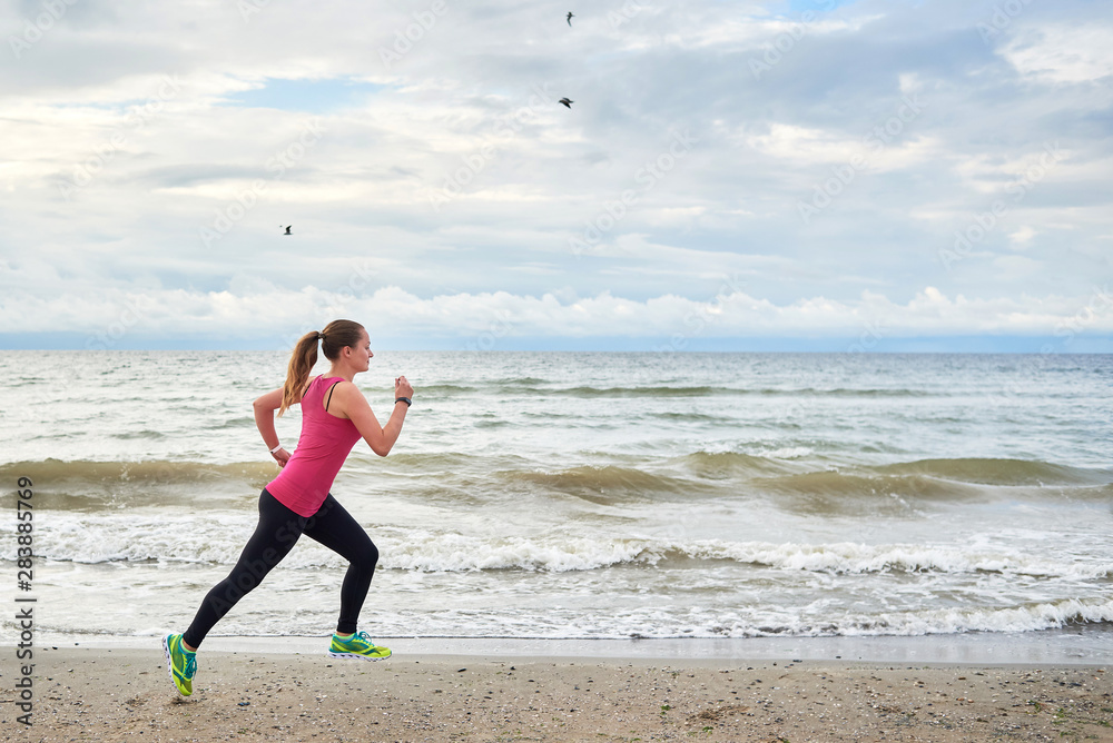Young fitness woman runner running at seaside, copy space. Girl working out on beach at summer morning, full length portrait. Side view. Healthy lifestyle concept