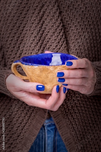 Beautiful female hands holding brown clay cup with blue enamel. Manicure with blue nail polish. Cozy concept