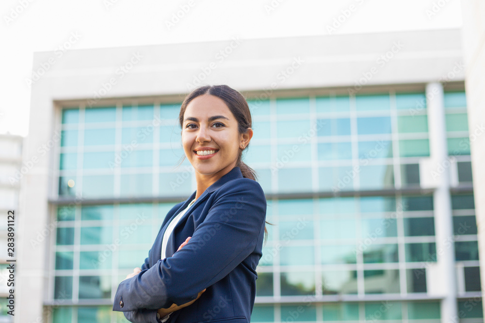 Happy successful young businesswoman posing outside. Beautiful Latin woman wearing formal jacket, standing near office building, looking at camera and smiling. Successful career concept Happy successful young businesswoman posing outside. Beautiful Latin woman wearing formal jacket, standing near office building, looking at camera and smiling. Successful career concept