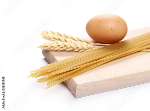 Raw spaghetti with wheat ears and egg on wooden chopping, cutting board isolated on white background