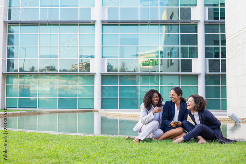 Happy female business colleagues enjoying work break outdoors. Women wearing office suits, sitting on grass near office building, chatting and laughing. Business and friendship concept © Mangostar