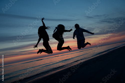 Three 3 women jumps into air and raises their hands up. Silhouette of three girls jumping near the sea, nice sunset and people enjoying near the sea.Happy girl rejoices summer.Women figure,silhouette
