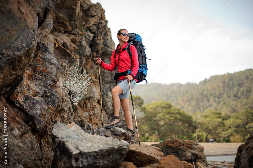 Smiling woman standing on the rocks with hiking backpack and walking sticks