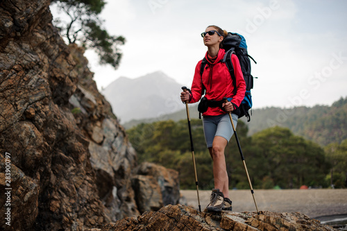 Slim fit girl in special wear standing with hiking backpack and sticks on the rock