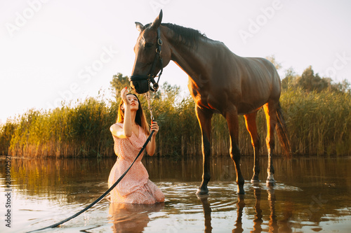 Woman sits next to the horse on the river at sunset.