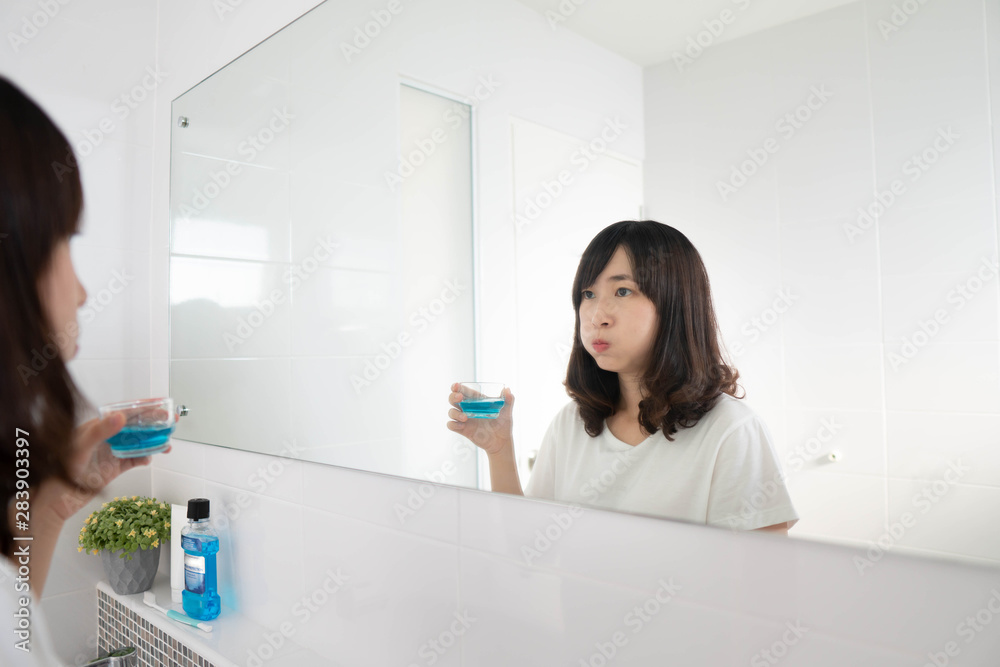 Young asian woman rinsing and gargling mouth with mouthwash after brushing her teeth in bathroom. Oral hygiene routine for freshness breath, prevent plaque and gum disease. Dental health care concept. Young asian woman rinsing and gargling mouth with mouthwash after brushing her teeth in bathroom. Oral hygiene routine for freshness breath, prevent plaque and gum disease. Dental health care concept.
