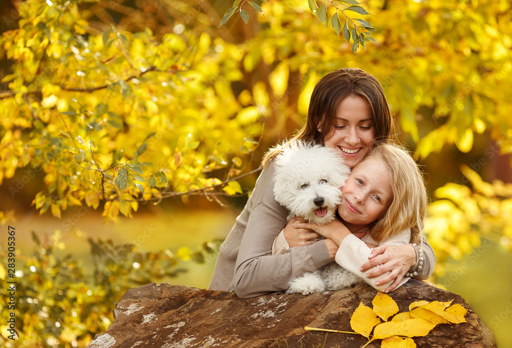Happy family mom and daughter with little white dog hug and laugh sunny autumn day on the background of yellow leaves Happy family mom and daughter with little white dog hug and laugh sunny autumn day on the background of yellow leaves