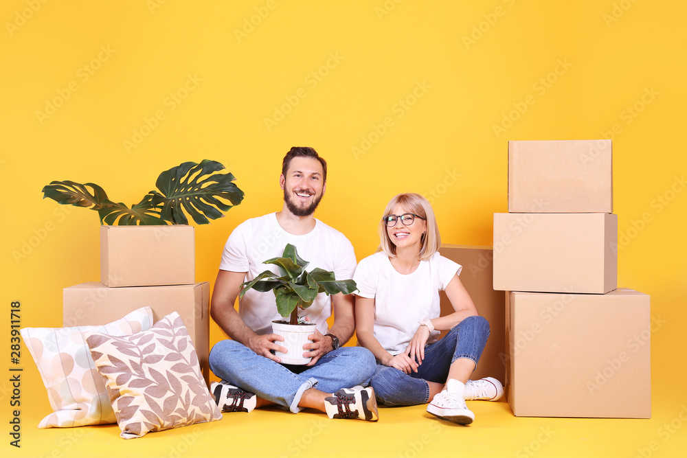 Young married couple moving in new apartment, sitting on the floor between many boxes. New home concept. Bearded man and blonde woman resting over yellow wall. Background, copy space, close up. Young married couple moving in new apartment, sitting on the floor between many boxes. New home concept. Bearded man and blonde woman resting over yellow wall. Background, copy space, close up.
