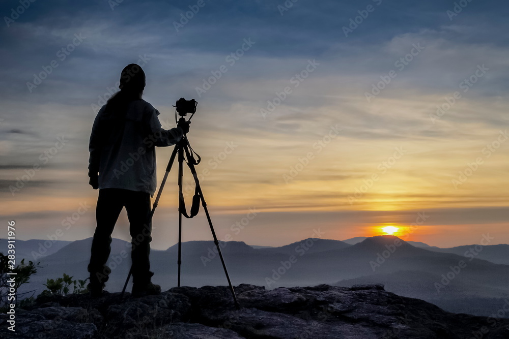 Silhouette a Photographer standing on top hill above Mekong river around with soft mist, mountains and cloudy sky background, sunrise at Cha Na Dai Cliff, Pha Taem National Park, Ubon, Thailand.
