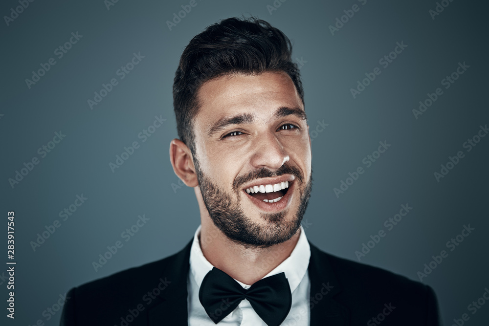Charming young man in bow tie looking at camera and smiling while standing against grey background Charming young man in bow tie looking at camera and smiling while standing against grey background