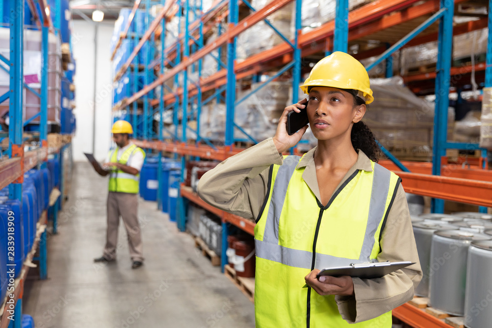 Female worker holding clipboard and talking on mobile phone in warehouse Female worker holding clipboard and talking on mobile phone in warehouse