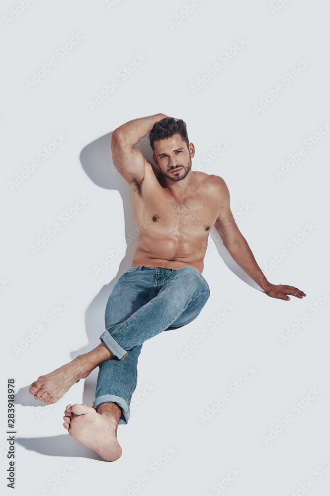 Handsome young shirtless man looking at camera while sitting against grey background Handsome young shirtless man looking at camera while sitting against grey background
