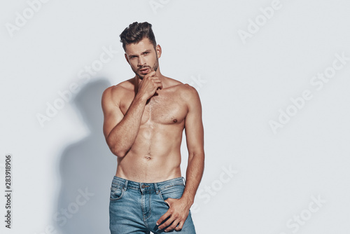 Handsome young shirtless man looking at camera and keeping hand on chin while standing against grey background