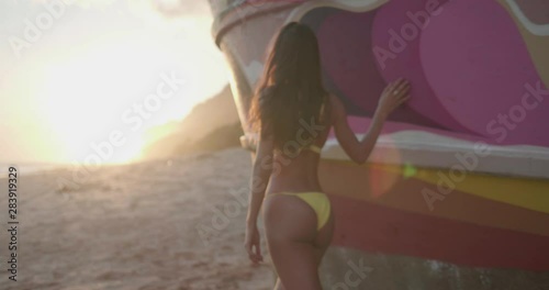 Back view of beautiful tanned fit happy woman in yellow bikini walking near the shipwreck at the beach during sunset - video in slow motion