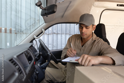 Delivery man with cardboard boxes writing on clipboard in front seat of car outside the warehouse