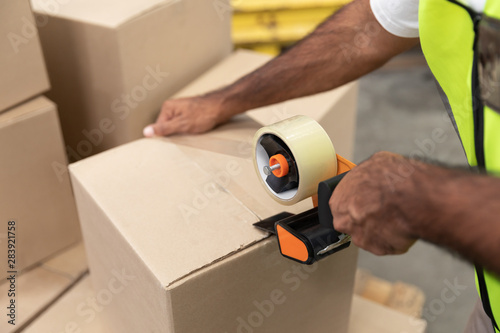 Male worker packing cardboard box with tape gun dispenser in warehouse photo