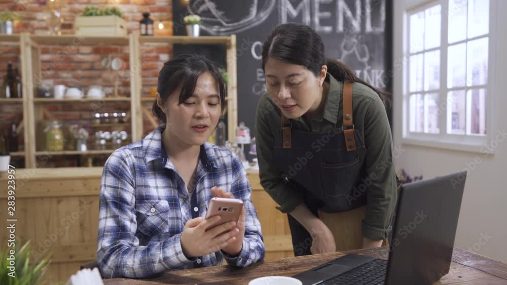 female barista chatting with girl guest in cafe store. asian chinese woman customer sitting at wooden table with laptop and showing waitress cellphone screen. Occupation people and service concept. female barista chatting with girl guest in cafe store. asian chinese woman customer sitting at wooden table with laptop and showing waitress cellphone screen. Occupation people and service concept.