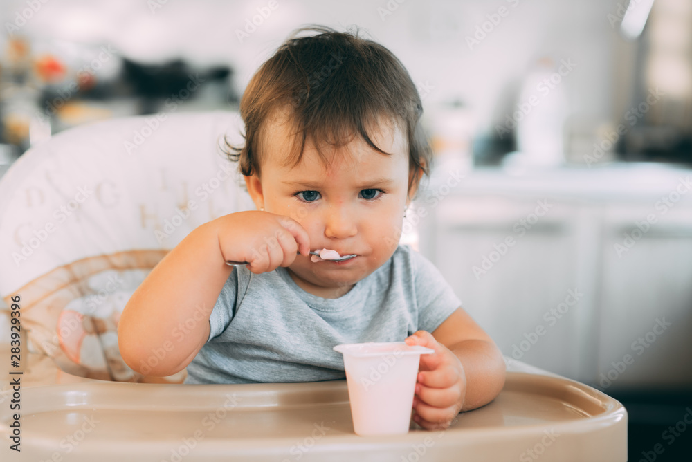Cute, little girl eating yogurt in the kitchen during the day in a high chair Cute, little girl eating yogurt in the kitchen during the day in a high chair