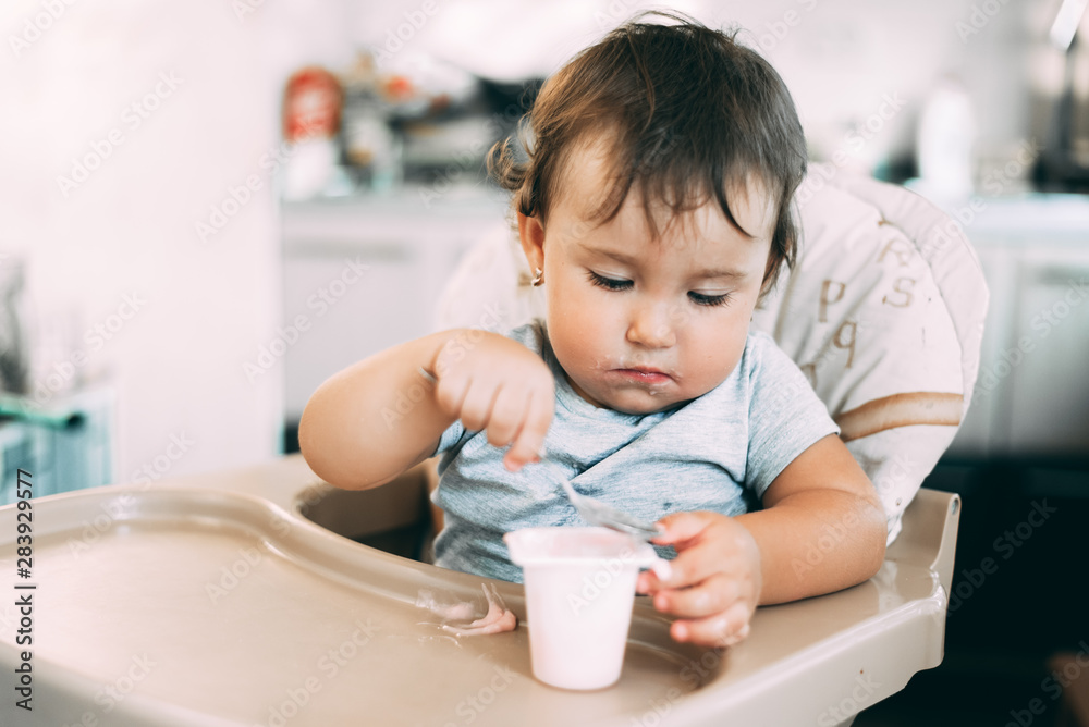Cute, little girl eating yogurt in the kitchen during the day in a high chair Cute, little girl eating yogurt in the kitchen during the day in a high chair