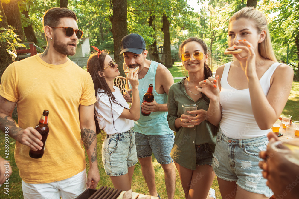 Group of happy friends having beer and barbecue party at sunny day. Resting together outdoor in a forest glade or backyard. Celebrating and relaxing, laughting. Summer lifestyle, friendship concept.
