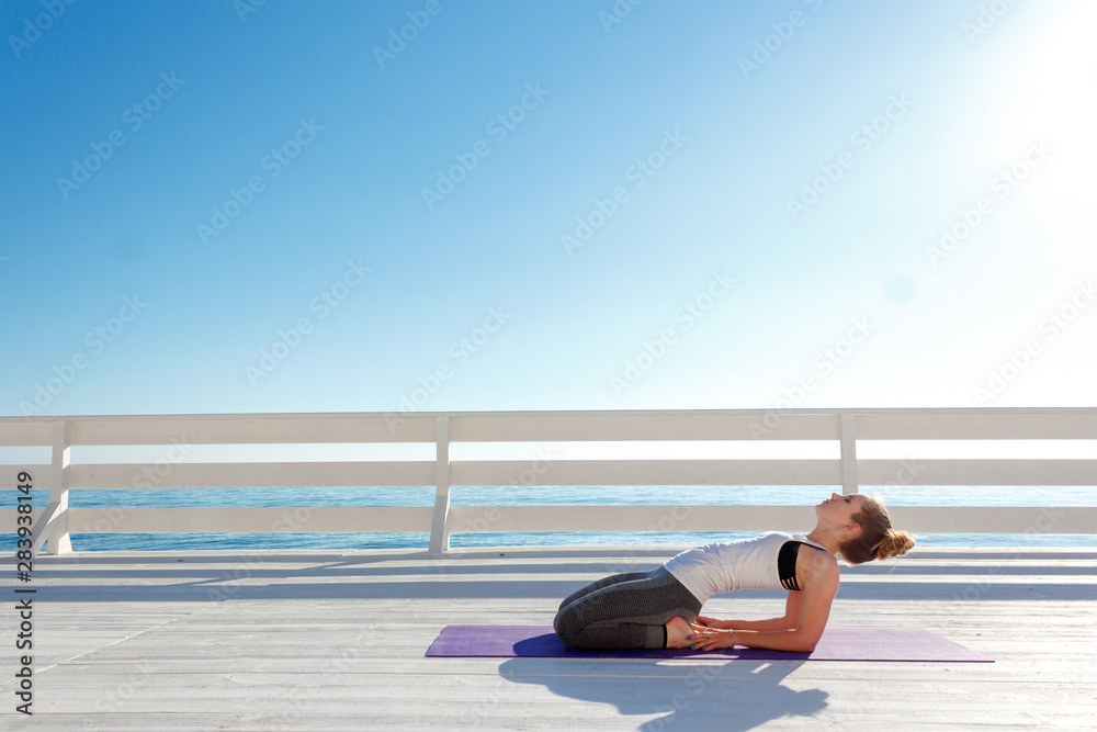 Fototapeta premium Young slim woman practicing yoga outdoors at white wooden seafront