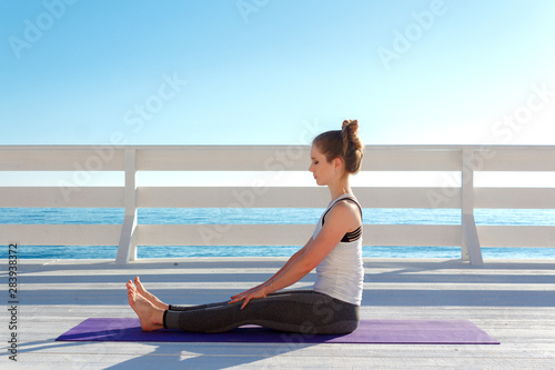Woman practicing yoga outdoors at white wooden seafront. Girl sits with straight legs and back © yolya_ilyasova