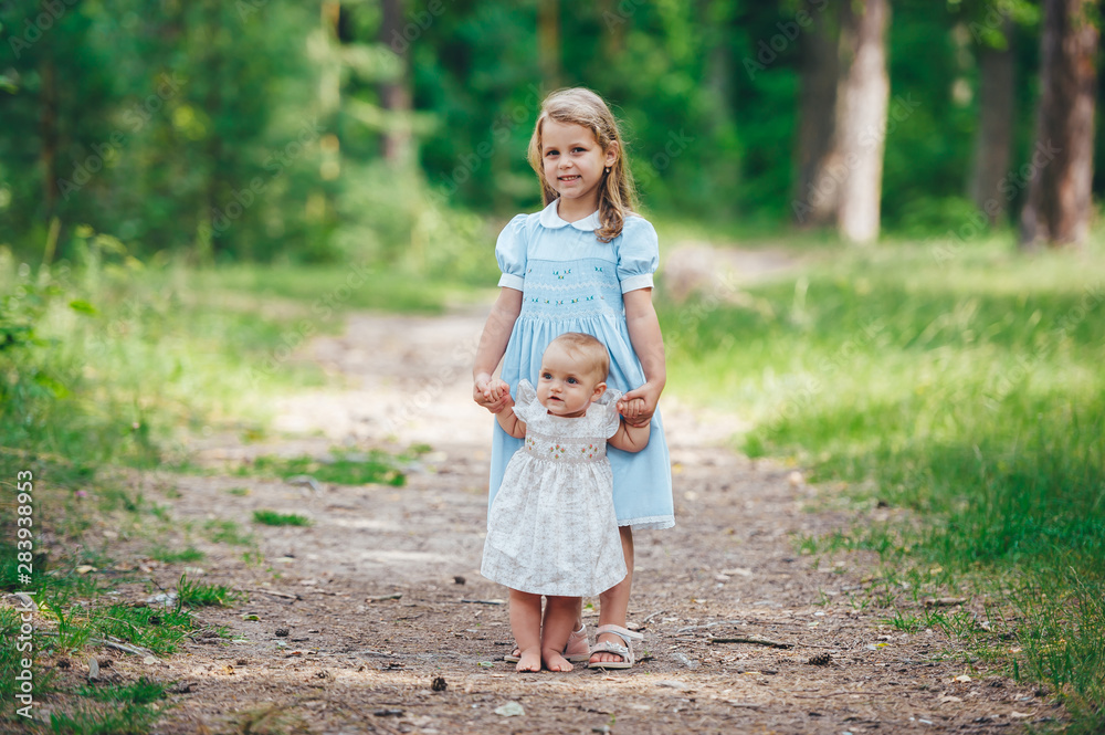 Two cute young sisters playing in sunny summer park and holding hands.